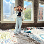 Artist Mary Ball standing with her arms behind her head in her studio, wearing the 'Seasons of Change' Shirt in blue open with jeans. 