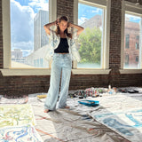 Artist Mary Ball standing with her arms behind her head in her studio, wearing the 'Seasons of Change' Shirt in blue open with jeans. 