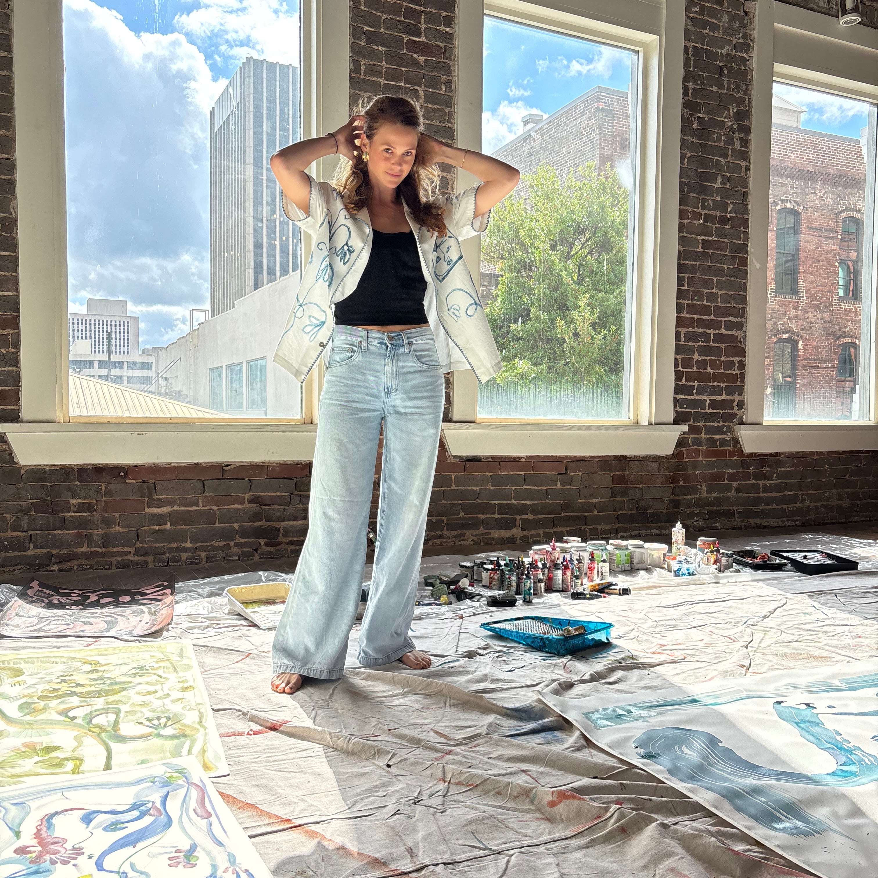Artist Mary Ball standing with her arms behind her head in her studio, wearing the 'Seasons of Change' Shirt in blue open with jeans. 
