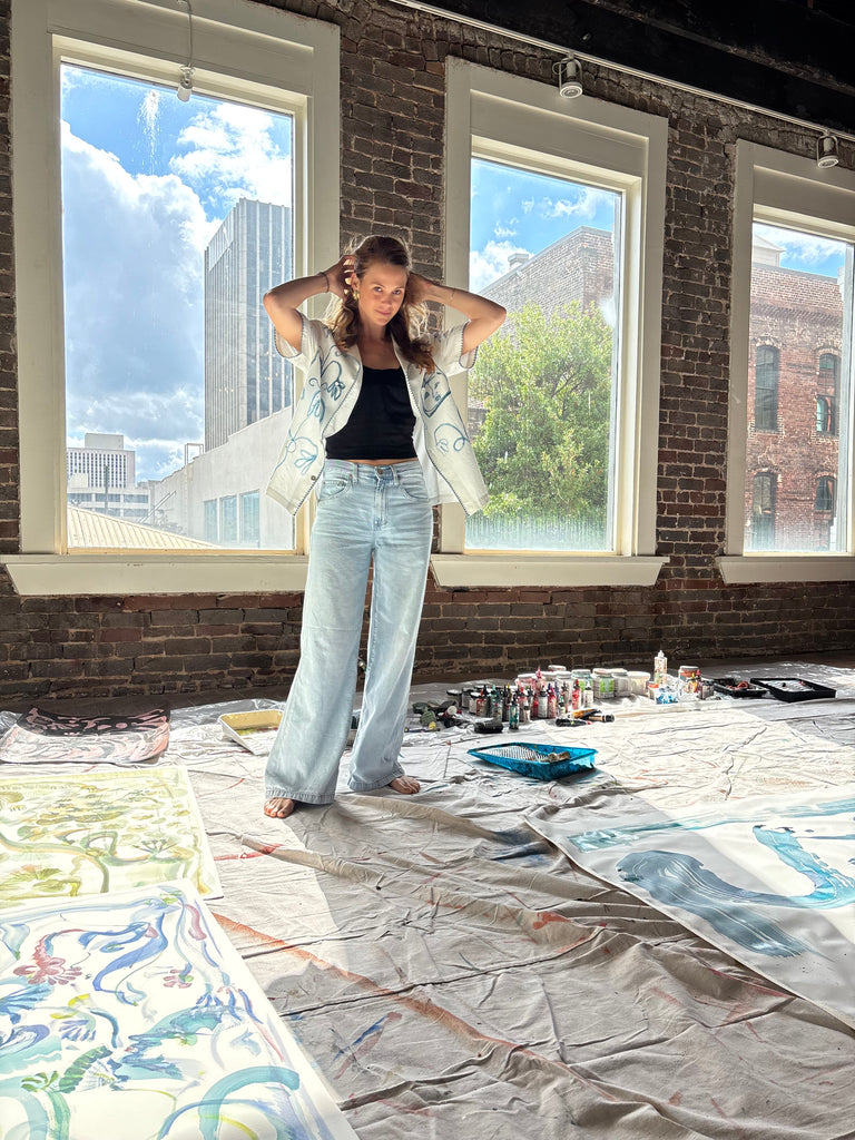 Artist Mary Ball standing with her arms behind her head in her studio, wearing the 'Seasons of Change' Shirt in blue open with jeans. 