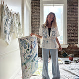 Artist Mary Ball holding one of her pieces, while wearing the 'Seasons of Change' Shirt in Blue with a pair of jeans. She is holding a paintbrush in her other hand!