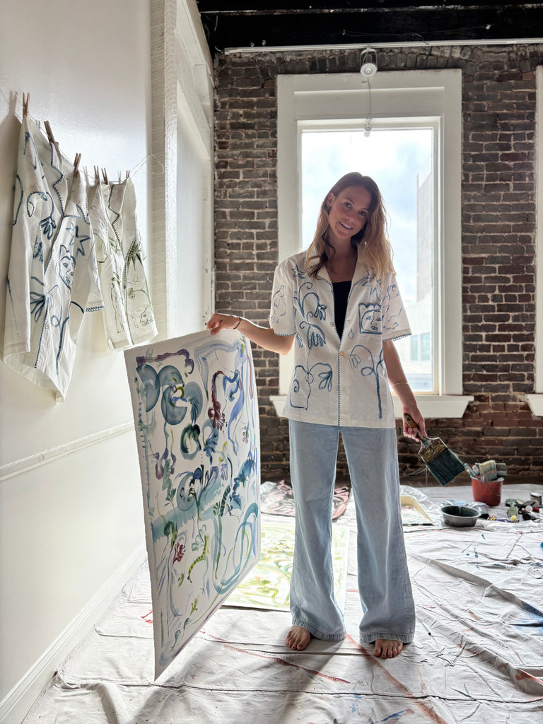 Artist Mary Ball holding one of her pieces, while wearing the 'Seasons of Change' Shirt in Blue with a pair of jeans. She is holding a paintbrush in her other hand!