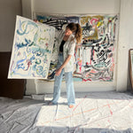Image of artist, Mary Ball, holding up one of her pieces while wearing the green 'Seasons of Change' Shirt. She is standing in front of a large canvas painting of hers, as well.