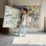 Image of artist, Mary Ball, holding up one of her pieces while wearing the green 'Seasons of Change' Shirt. She is standing in front of a large canvas painting of hers, as well.