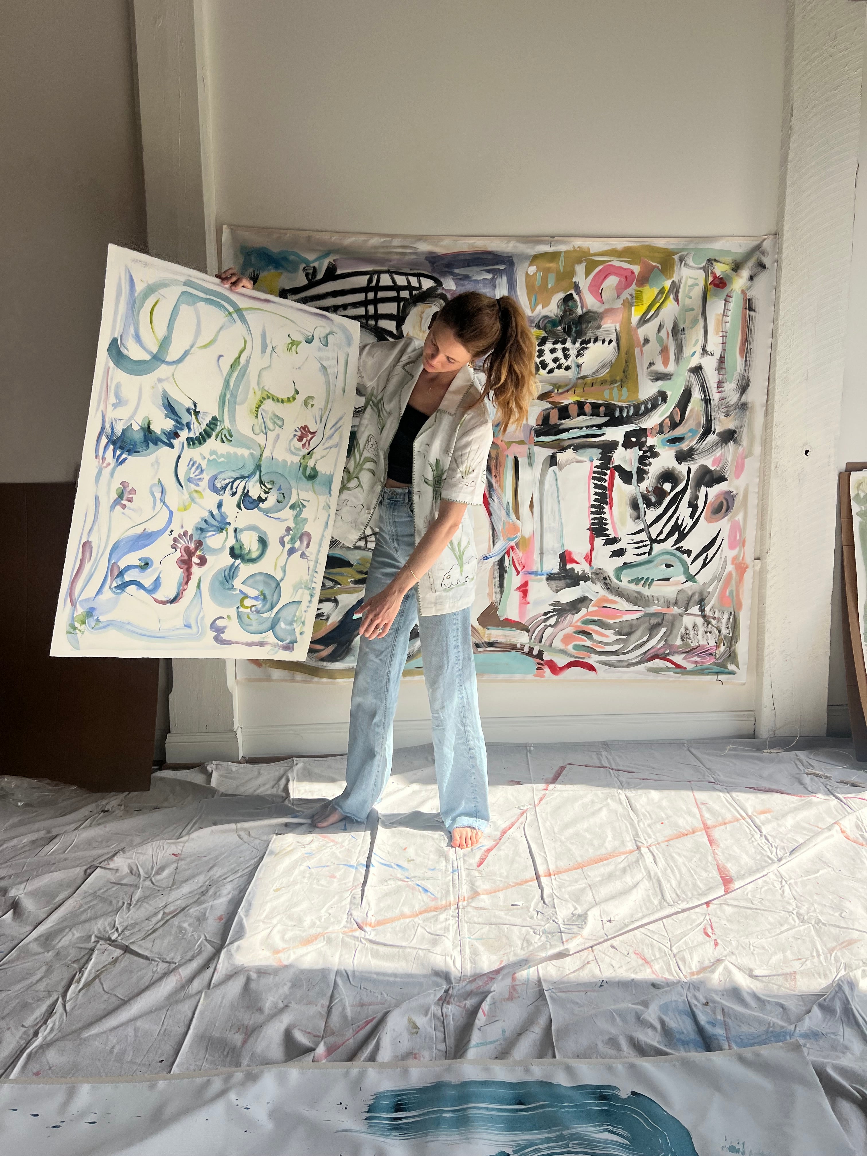 Image of artist, Mary Ball, holding up one of her pieces while wearing the green 'Seasons of Change' Shirt. She is standing in front of a large canvas painting of hers, as well.