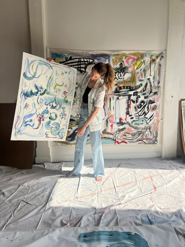 Image of artist, Mary Ball, holding up one of her pieces while wearing the green 'Seasons of Change' Shirt. She is standing in front of a large canvas painting of hers, as well.