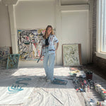 Artist Mary Ball standing in her studio with a paintbrush in hand, wearing the brown and blue abstract shirt she designed with Tombolo. 
