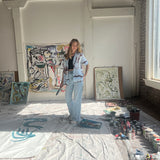 Artist Mary Ball standing in her studio with a paintbrush in hand, wearing the brown and blue abstract shirt she designed with Tombolo. 