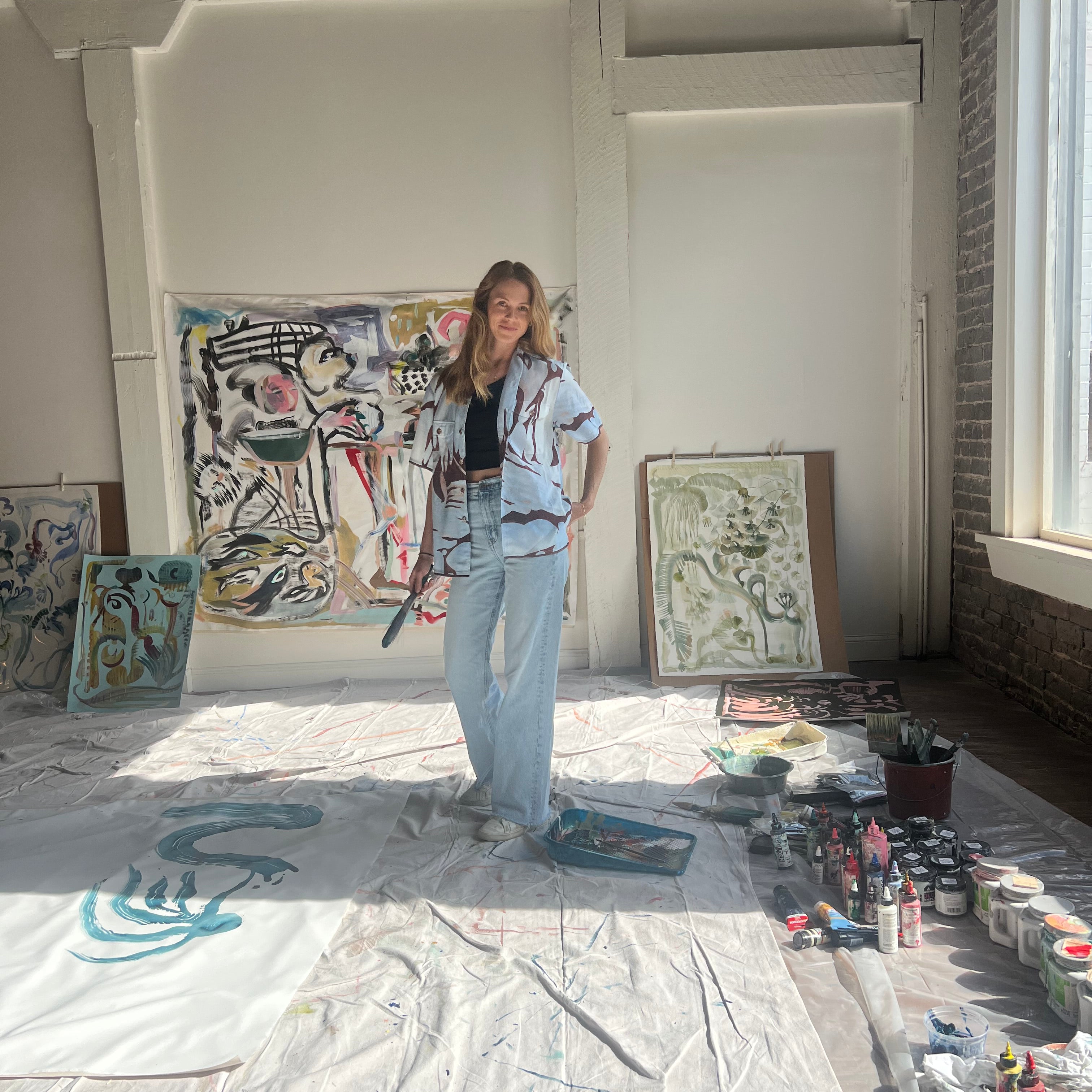 Artist Mary Ball standing in her studio with a paintbrush in hand, wearing the brown and blue abstract shirt she designed with Tombolo. 