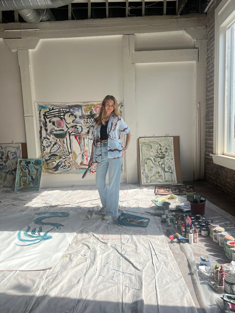 Artist Mary Ball standing in her studio with a paintbrush in hand, wearing the brown and blue abstract shirt she designed with Tombolo. 