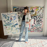 Image of artist, Mary Ball, holding up one of her pieces while wearing the green 'Seasons of Change' Shirt. She is standing in front of a large canvas painting of hers, as well.