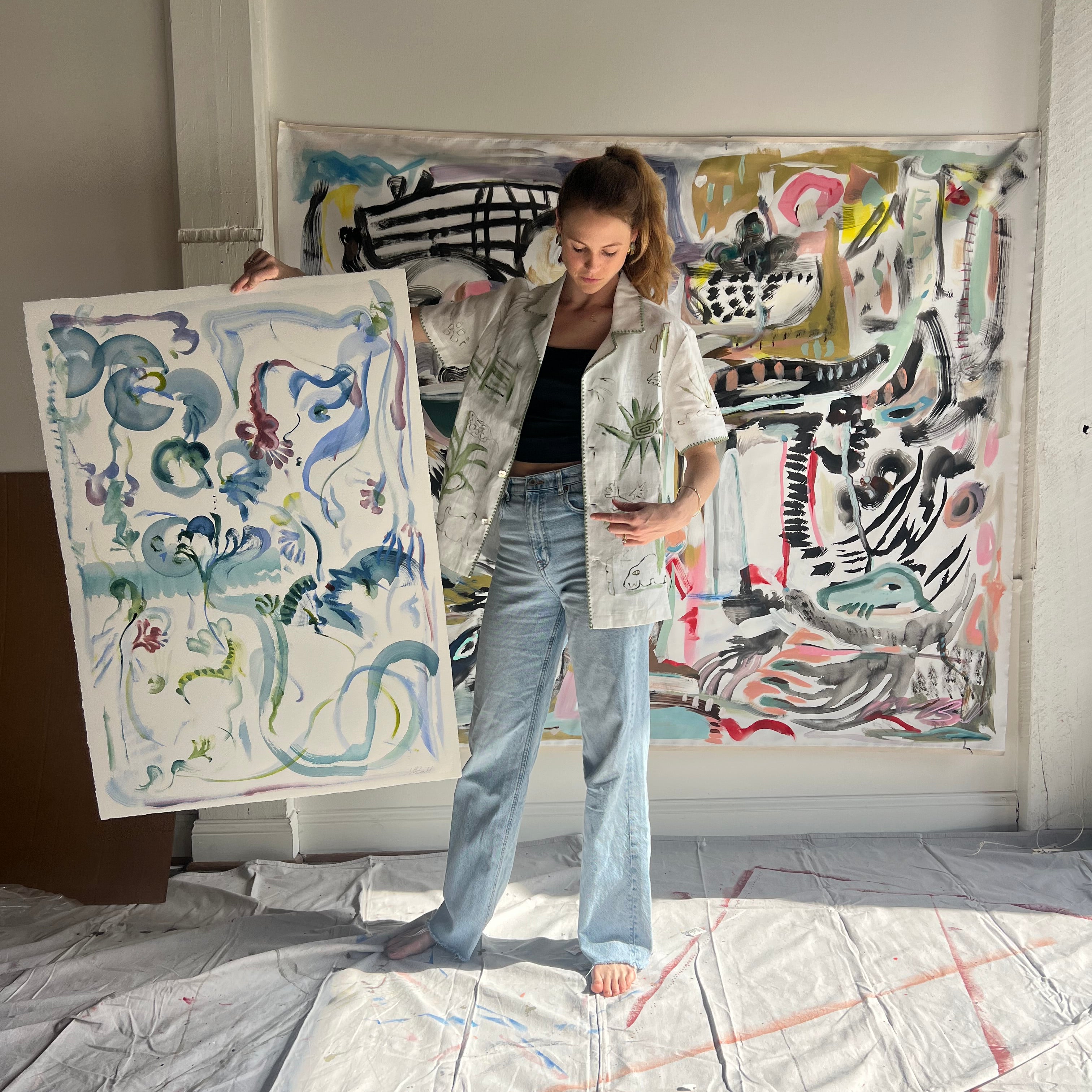 Image of artist, Mary Ball, holding up one of her pieces while wearing the green 'Seasons of Change' Shirt. She is standing in front of a large canvas painting of hers, as well.