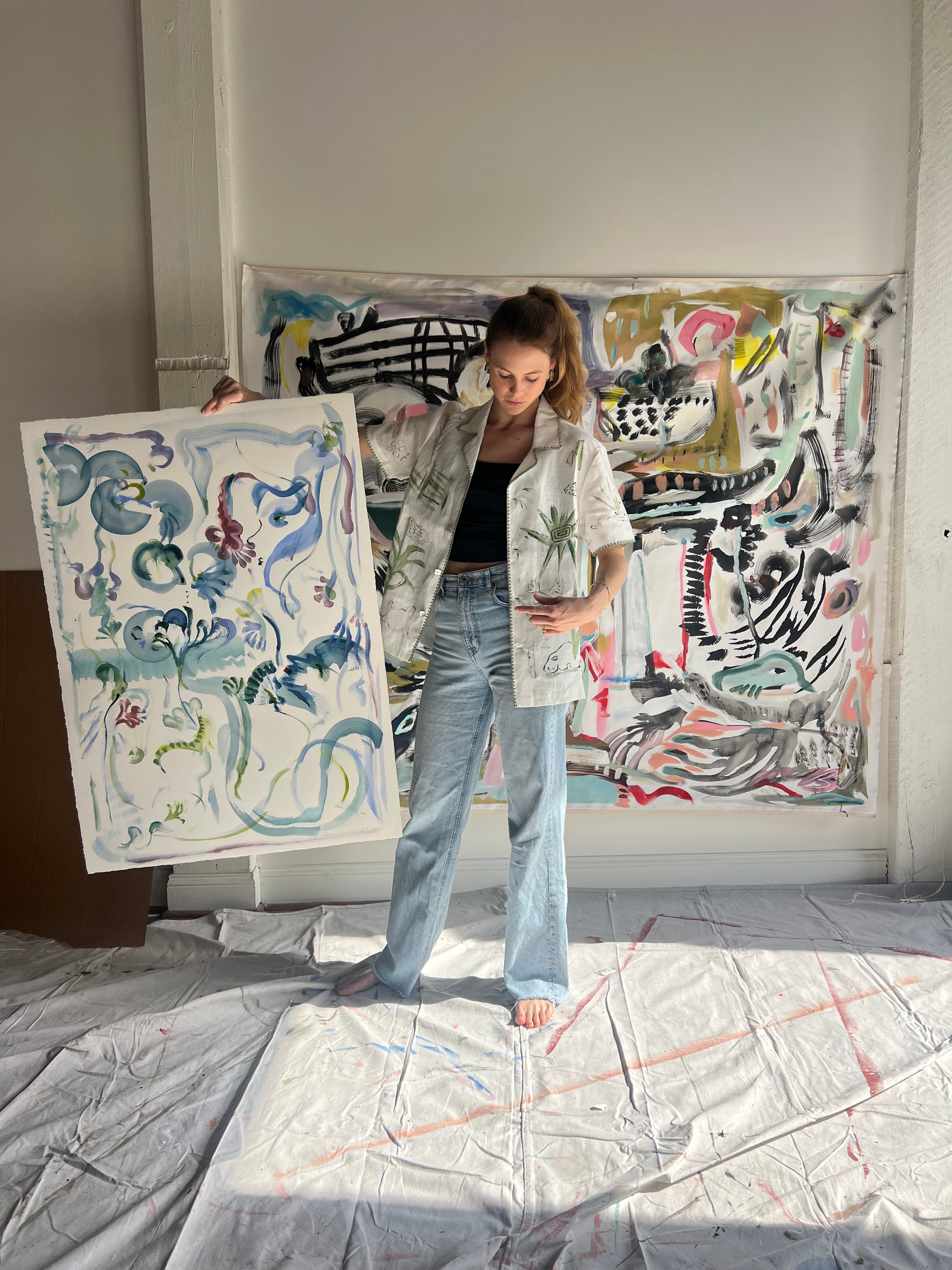Image of artist, Mary Ball, holding up one of her pieces while wearing the green 'Seasons of Change' Shirt. She is standing in front of a large canvas painting of hers, as well.