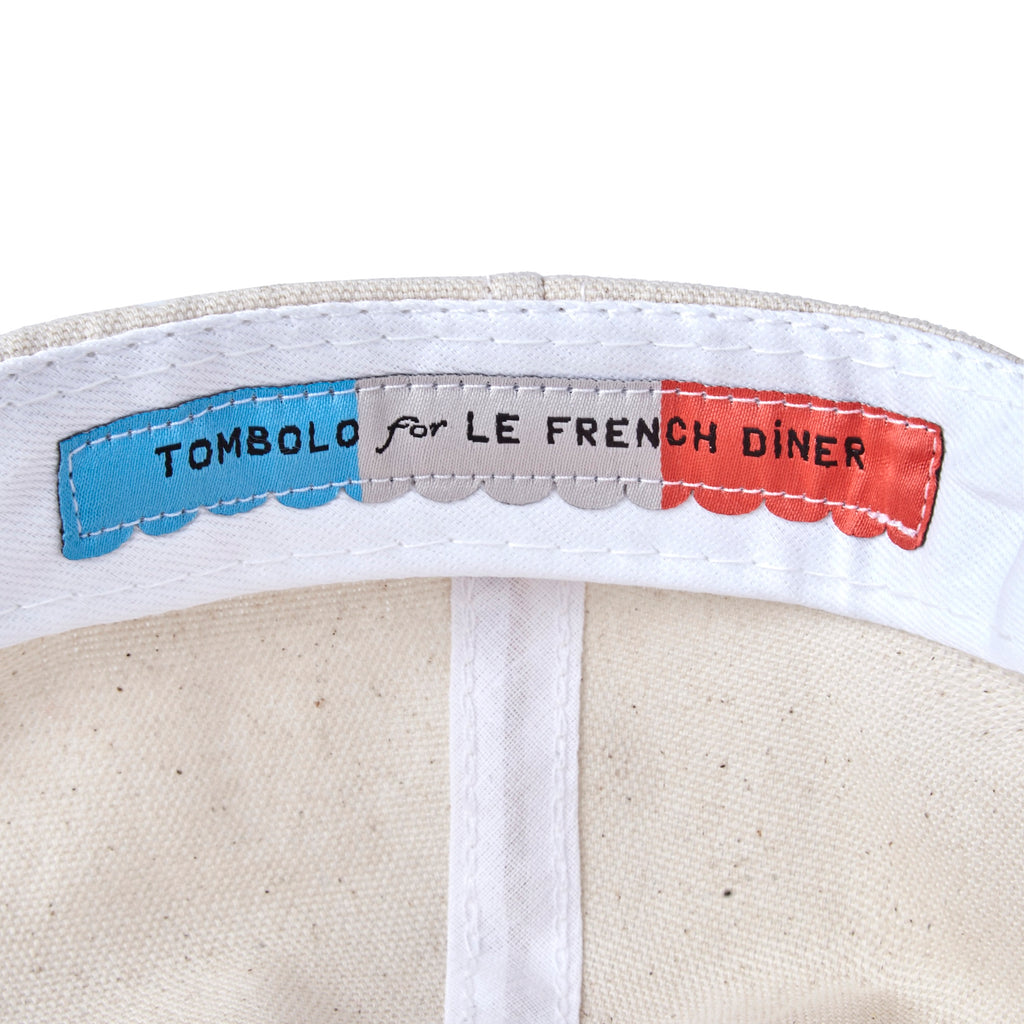 Close-up view of the inside label of a cap. The label features blue, gray, and red stitched panels reading “TOMBOLO for LE FRENCH DINER.”
