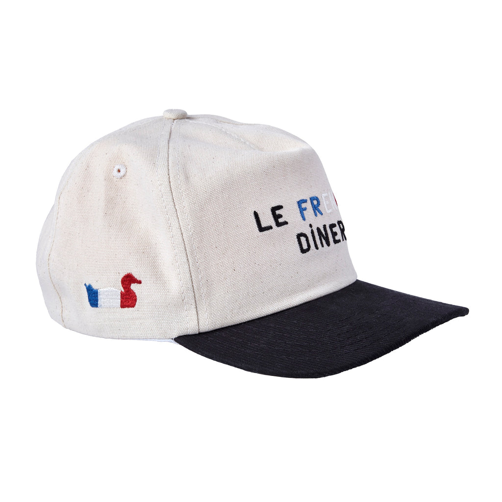 A beige canvas baseball cap with a black brim, shown from a side angle. The front is embroidered with the words “LE FRENCH DINER,” and the side features a small red duck next to a blue and white flag motif.