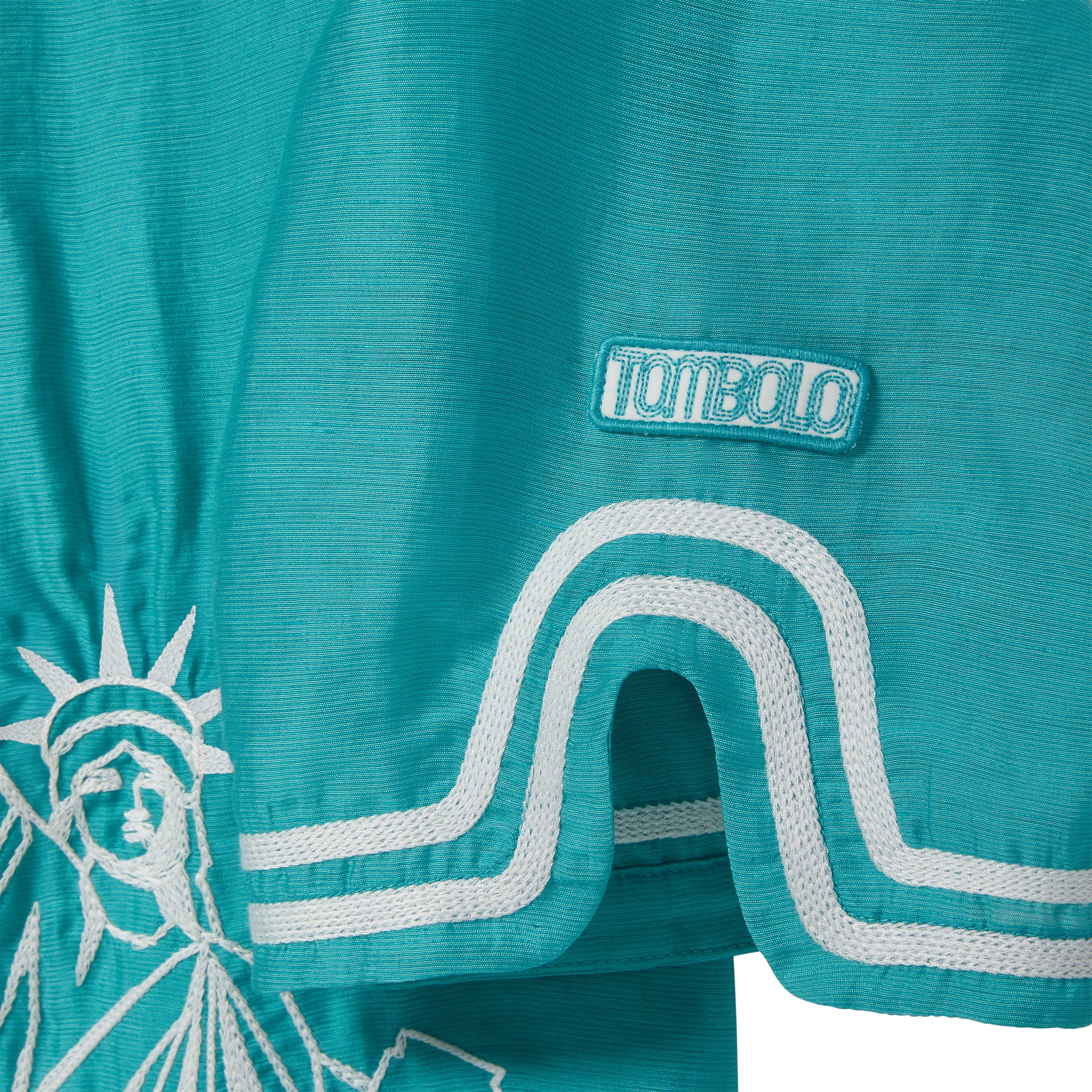 Teal blue shirt with white embroidery and white 'Tombolo' logo patch.