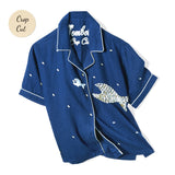 Front-facing product image of the 'Teamwork' Cabana ~ Crop Cut made of 60% Tencel and 40% Linen in a predominately navy coloring with a hand appliqué of a large fish chasing a smaller fish, fish-shaped agoya shell buttons serve as functional buttons on the placket and are scattered across the body, on the wearer's left panel, the fish-shaped buttons have been carefully assembled into the shape of a larger fish, chain-stitched interior label, and light-blue piping