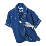 Front-facing product image of the 'Teamwork' Cabana ~ Crop Cut made of 60% Tencel and 40% Linen in a predominately navy coloring with a hand appliqué of a large fish chasing a smaller fish, fish-shaped agoya shell buttons serve as functional buttons on the placket and are scattered across the body, on the wearer's left panel, the fish-shaped buttons have been carefully assembled into the shape of a larger fish, chain-stitched interior label, and light-blue piping