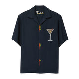 A navy blue short-sleeve button-down shirt with playful detailing: embroidered olive skewers act as buttons down the front, and a white martini glass with an olive garnish is embroidered on the chest. 