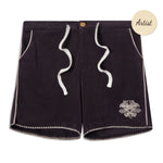 A pair of black terry cloth drawstring shorts with white zigzag trim along the hem and side seams. The shorts feature white rope-style drawstrings at the front and a small embroidered design resembling a stylized face or totem near the left hem. The waistband is elastic with a single wooden button at the top. A circular "Artist" badge appears in the top right corner of the image.