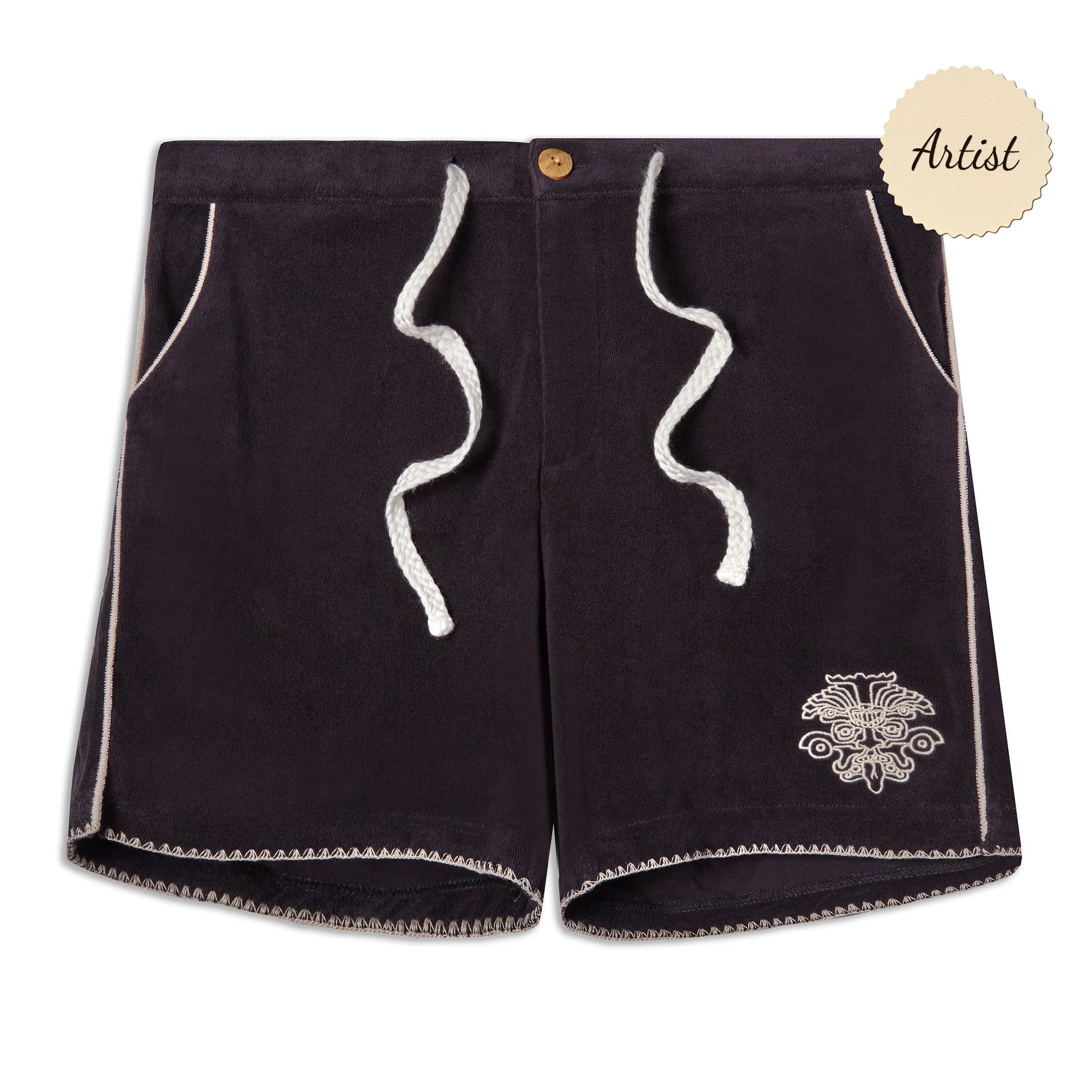 A pair of black terry cloth drawstring shorts with white zigzag trim along the hem and side seams. The shorts feature white rope-style drawstrings at the front and a small embroidered design resembling a stylized face or totem near the left hem. The waistband is elastic with a single wooden button at the top. A circular "Artist" badge appears in the top right corner of the image.