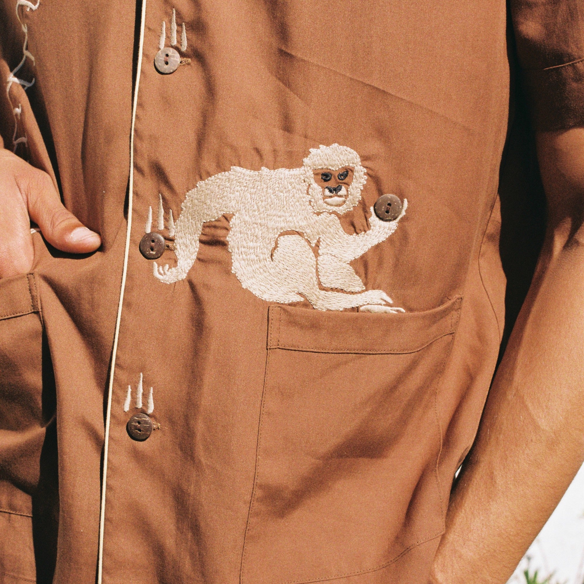 close up of male model wearing our breathable, lightweight Tencel Cabana Shirt featuring a carefully embroidered monkey catching a falling coconut  from a palm tree with buttons standing in for coconuts.