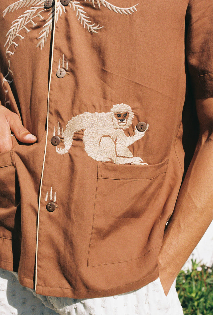 close up of male model wearing our breathable, lightweight Tencel Cabana Shirt featuring a carefully embroidered monkey catching a falling coconut  from a palm tree with buttons standing in for coconuts.