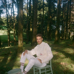 Man sits on chair with feet resting against a stool in the middle of forest