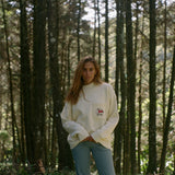 Woman subtly lifts up the bottom of sweater amidst a backdrop of tall trees
