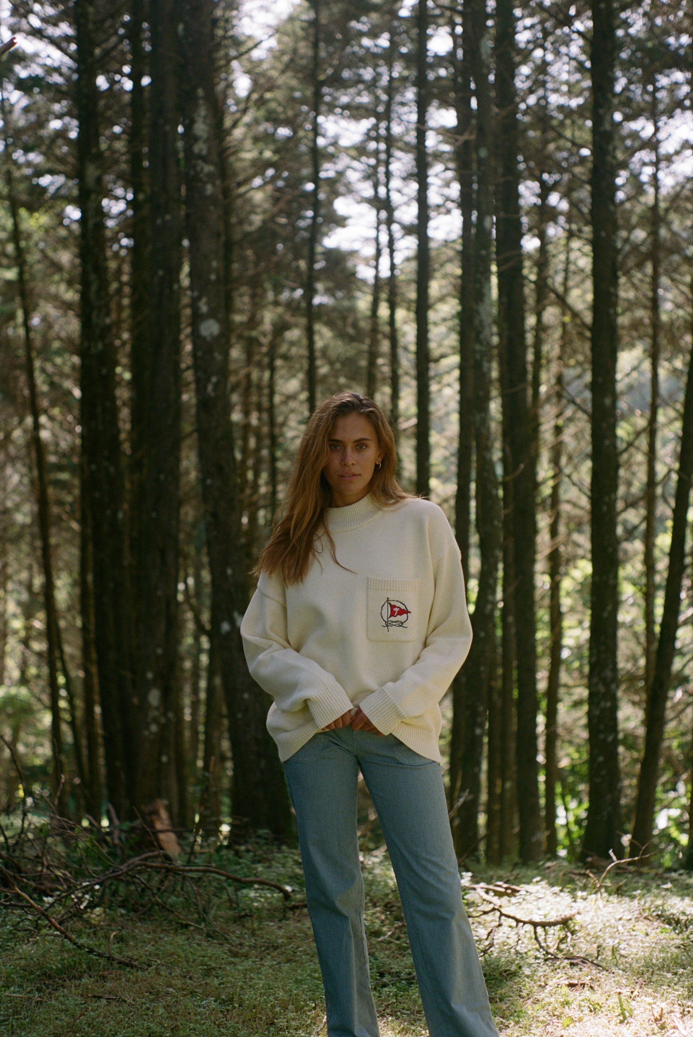 Woman subtly lifts up the bottom of sweater amidst a backdrop of tall trees