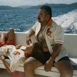 Two people relaxing on a moving boat with ocean and island views in the background, both wearing the 'Crouching Tiger" Cabana Shirt.