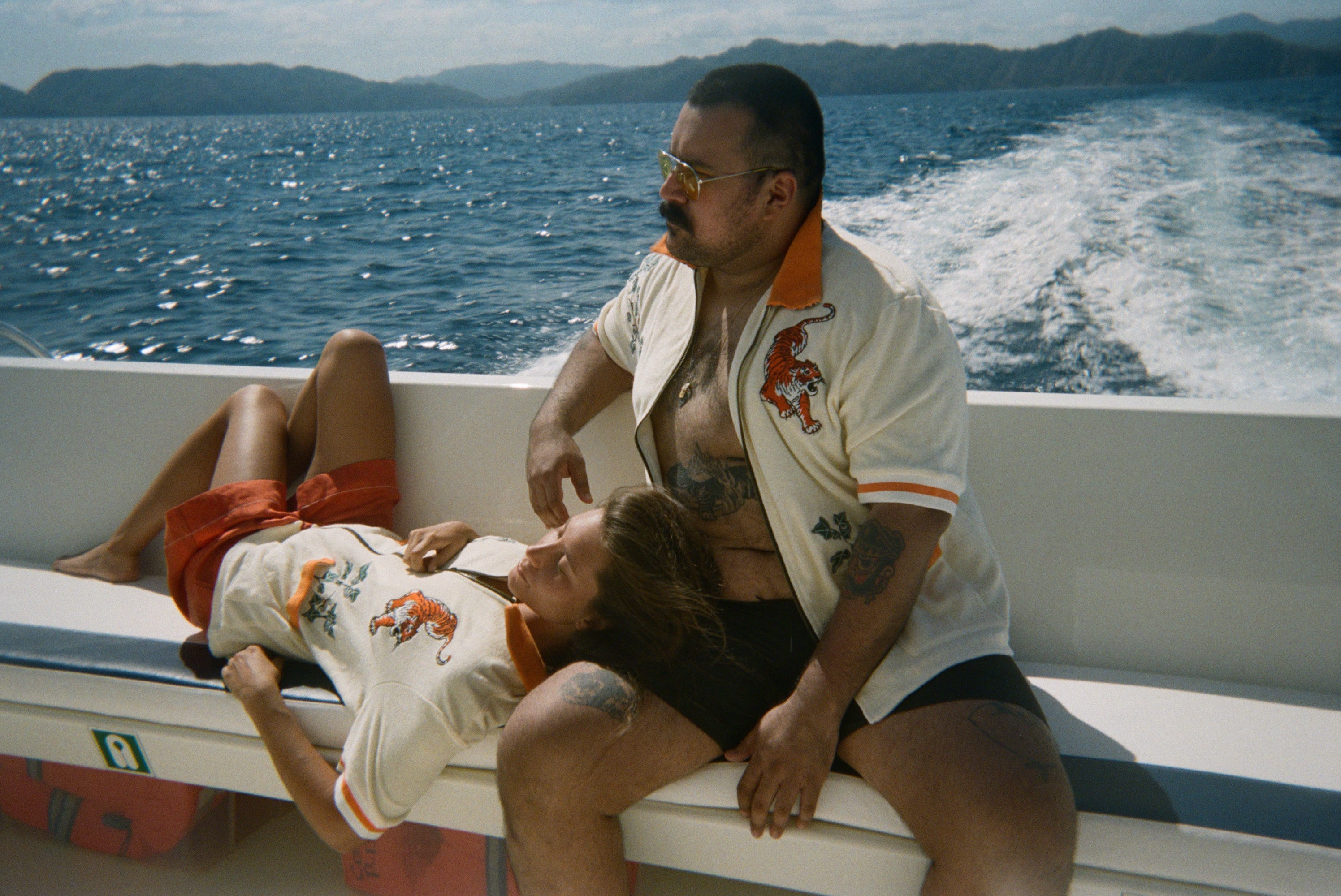 Two people relaxing on a moving boat with ocean and island views in the background, both wearing the 'Crouching Tiger" Cabana Shirt.