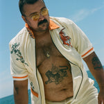 Man leaning forward on a boat under a clear blue sky, wearing the 'Crouching Tiger' Cabana opened. He has bold tiger tattoo on his stomach, wears yellow-tinted sunglasses, and sports a close-cropped haircut and mustache.