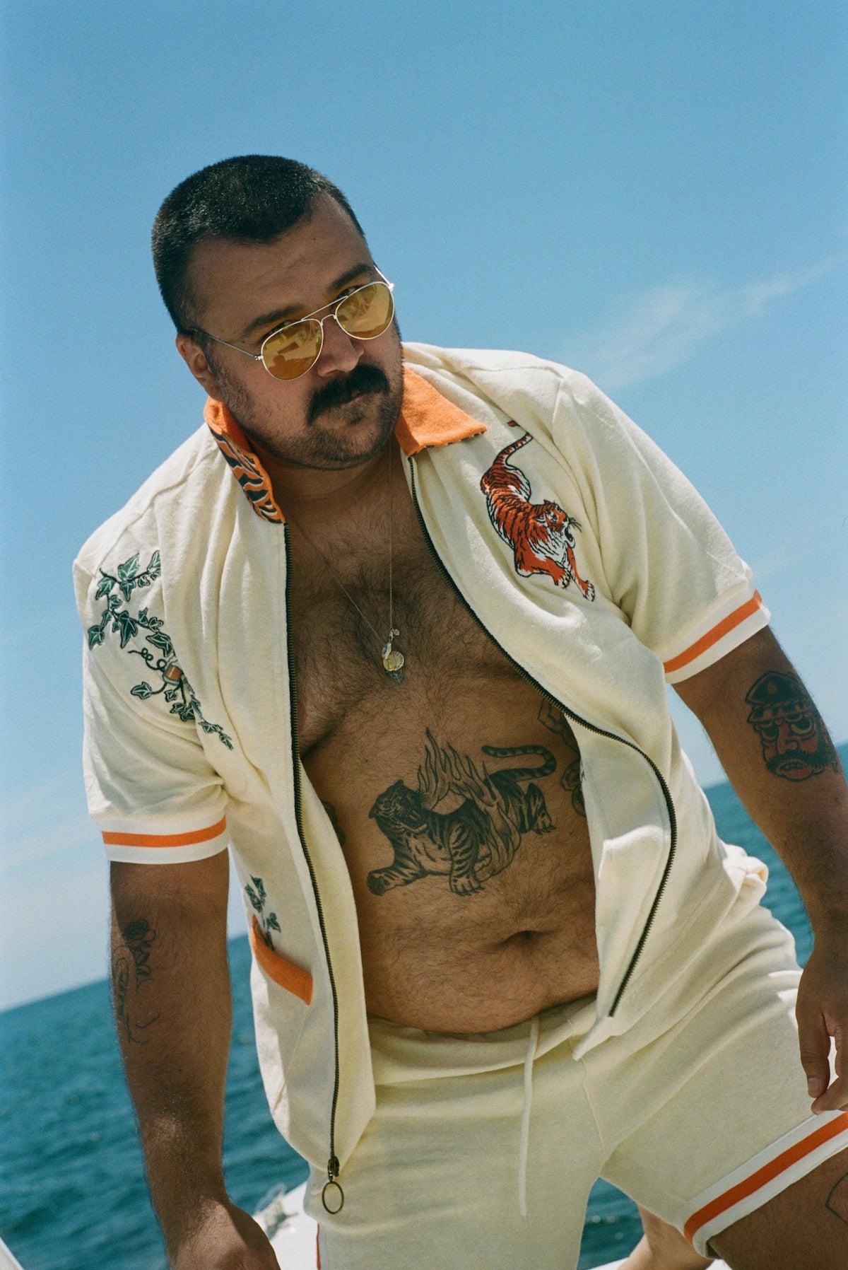 Man leaning forward on a boat under a clear blue sky, wearing the 'Crouching Tiger' Cabana opened. He has bold tiger tattoo on his stomach, wears yellow-tinted sunglasses, and sports a close-cropped haircut and mustache.