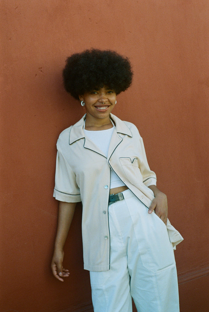 Female model wearing shirt with one button buttoned leaning on wall