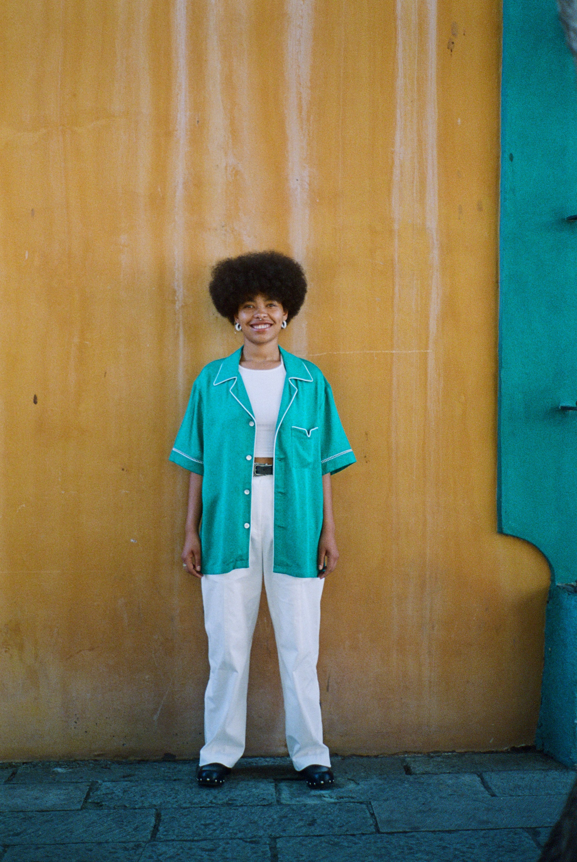 Female model wearing teal cabrisa over all white outfit standing against a wooden wall