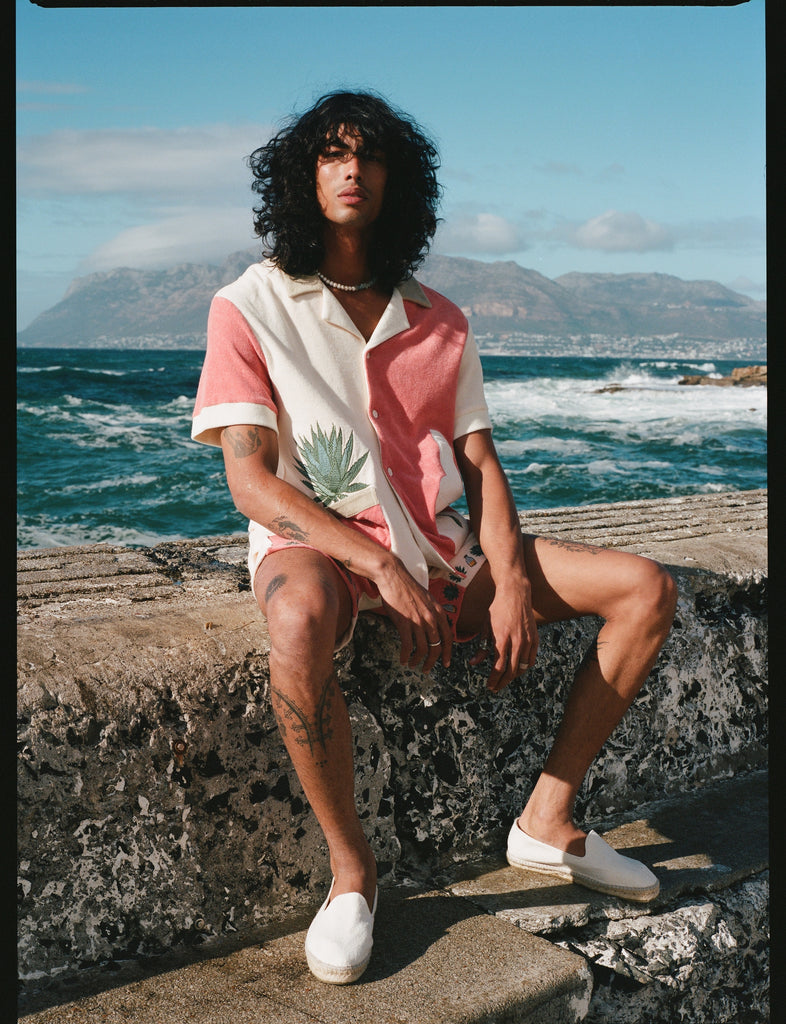 Male model sitting on a ledge by the sea wearing the full Gusanito set