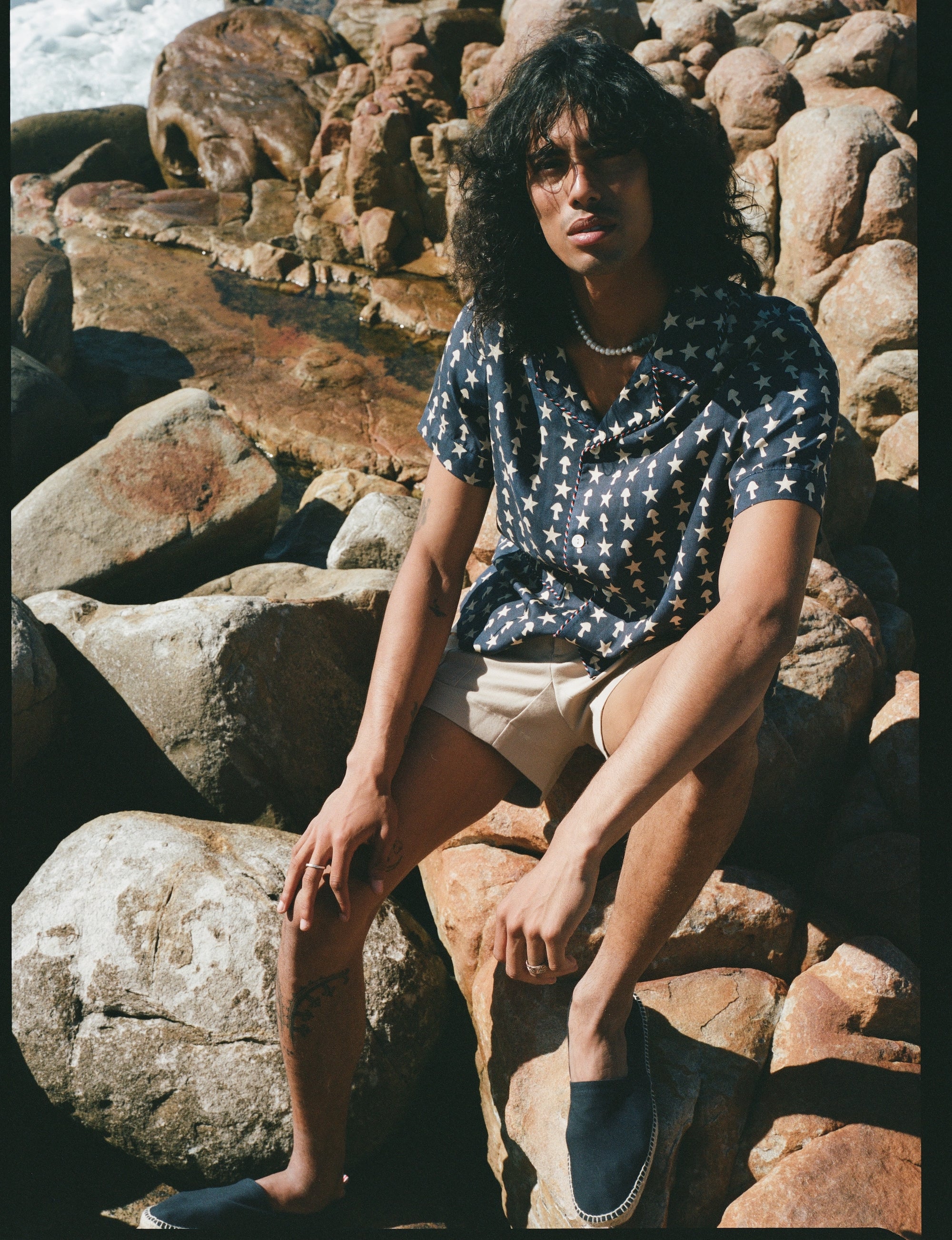 Male model wearing previous outfit sitting on a rocky beach
