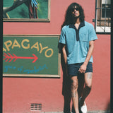 Male model leaning on pink wall wearing blue polo