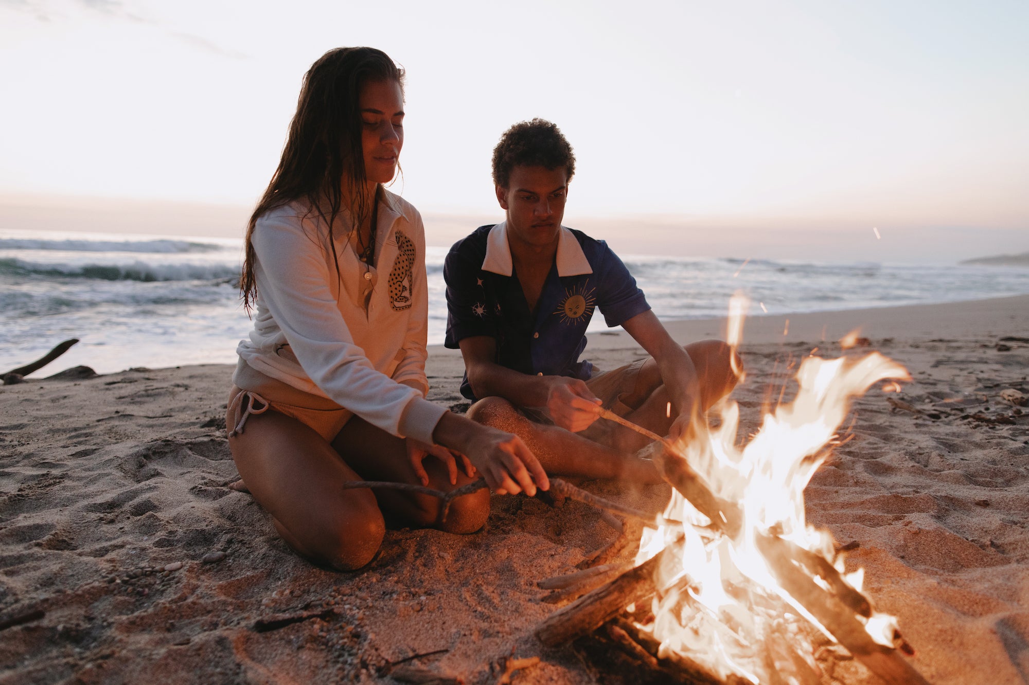 Female model wearing sun leopard cabana next to male model wearing rocket lobster cabana both putting sticks into a beachside fire