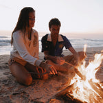 Female model wearing sun leopard cabana next to male model wearing rocket lobster cabana both putting sticks into a beachside fire