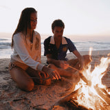 Female model wearing sun leopard cabana next to male model wearing rocket lobster cabana both putting sticks into a beachside fire