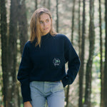Woman in the woods looks off camera wearing Mariner sweater