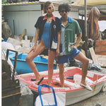 male and female models posing in boat with tote bag leaning against boat on sandy ground