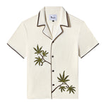 Front view of an off-white short-sleeve terrycloth cabana shirt with brown trim and dark brown buttons. Two embroidered green cannabis leaves with brown stems appear on the left chest and lower right side, creating a natural, asymmetric design. The collar and sleeve edges feature a zigzag brown stitch detail for added contrast.