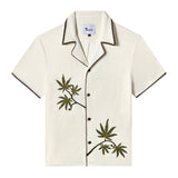 Front view of an off-white short-sleeve terrycloth cabana shirt with brown trim and dark brown buttons. Two embroidered green cannabis leaves with brown stems appear on the left chest and lower right side, creating a natural, asymmetric design. The collar and sleeve edges feature a zigzag brown stitch detail for added contrast.