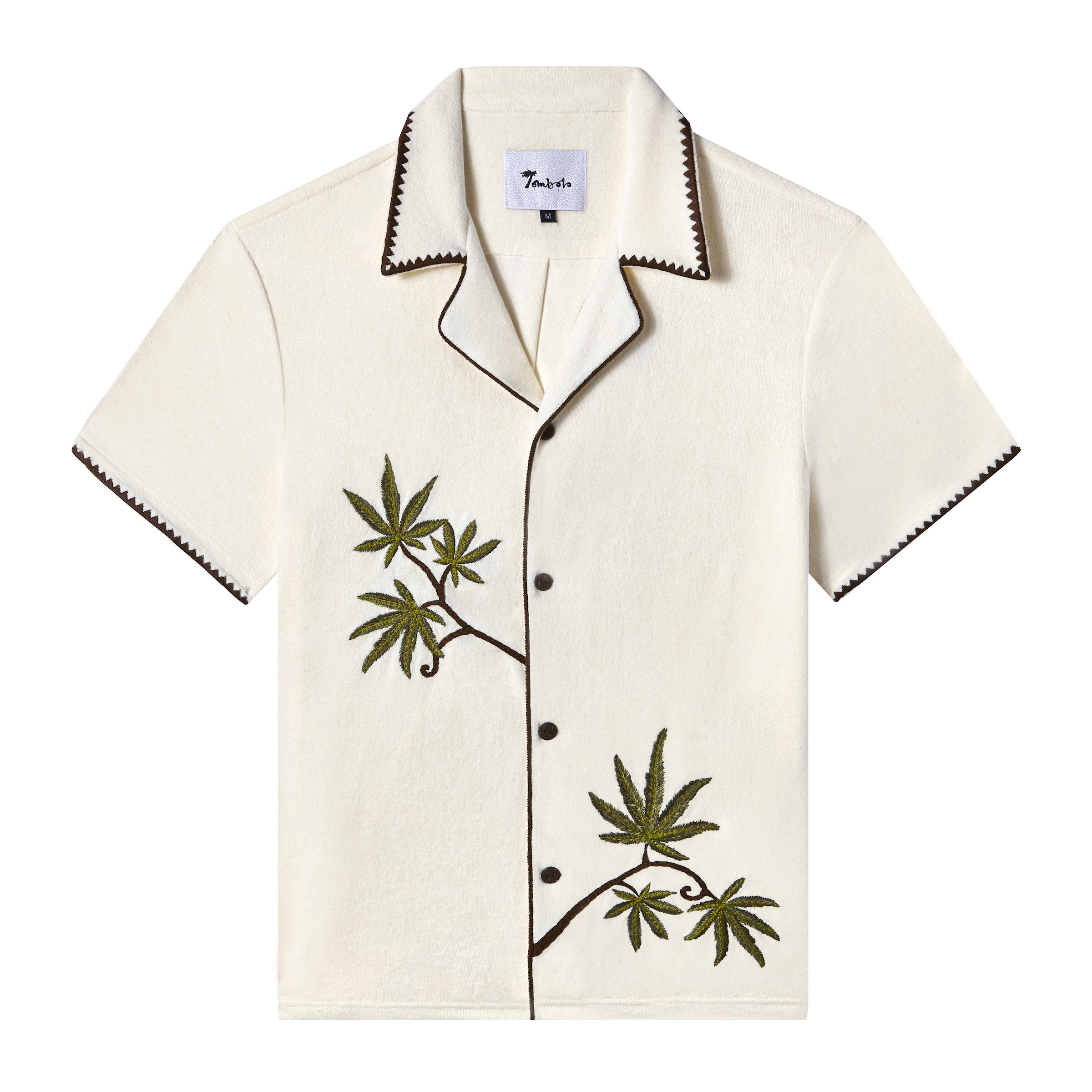 Front view of an off-white short-sleeve terrycloth cabana shirt with brown trim and dark brown buttons. Two embroidered green cannabis leaves with brown stems appear on the left chest and lower right side, creating a natural, asymmetric design. The collar and sleeve edges feature a zigzag brown stitch detail for added contrast.