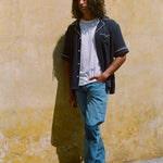 Male model wearing shirt unbuttoned over a white shirt and blue jeans against a wall