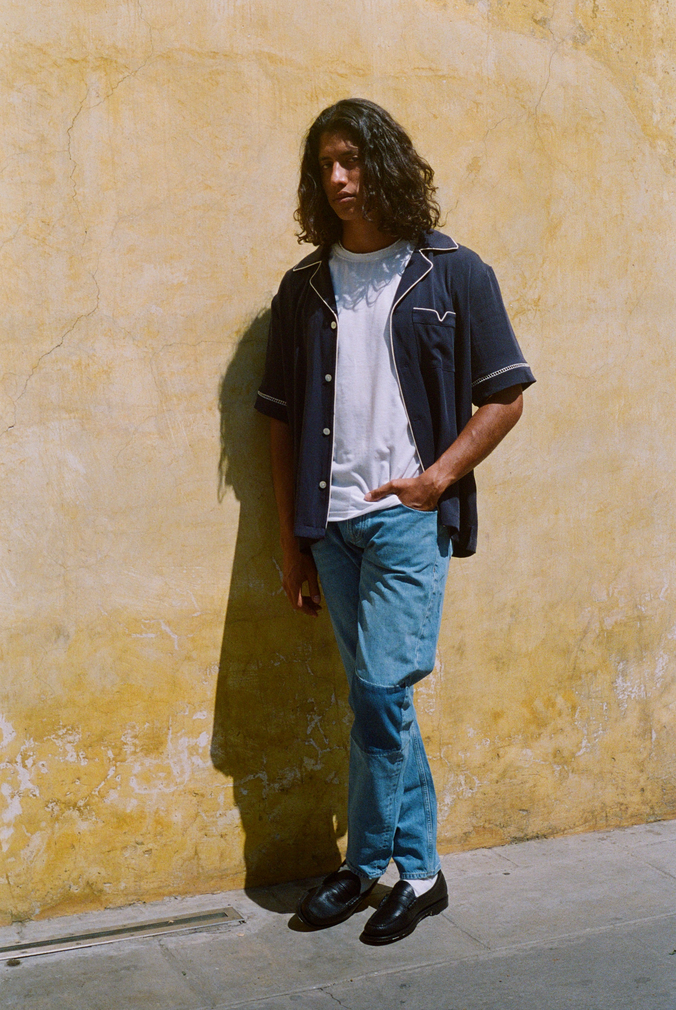 Male model wearing shirt unbuttoned over a white shirt and blue jeans against a wall