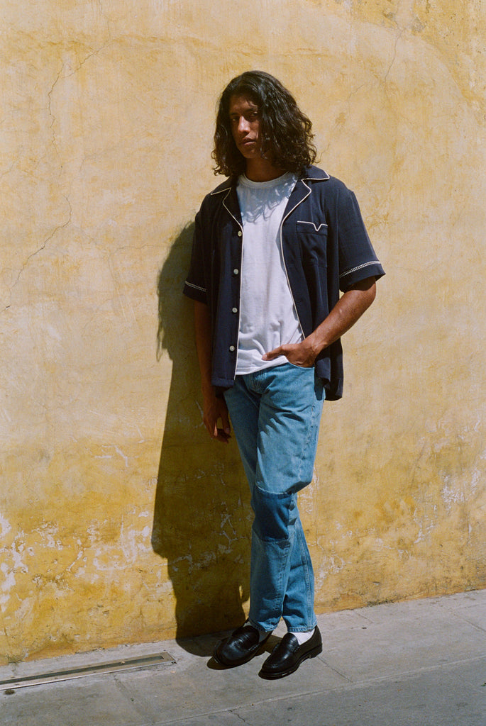 Male model wearing shirt unbuttoned over a white shirt and blue jeans against a wall